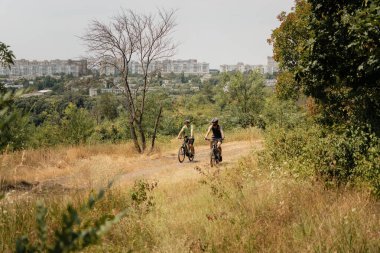 Two people riding bicycles on a dirt trail surrounded by trees and city view