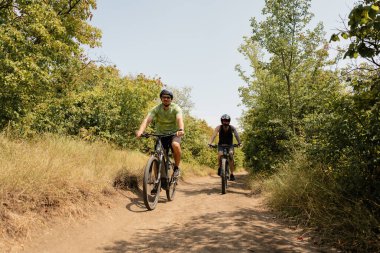 Two individuals cycling along a trail surrounded by nature on a bright day.