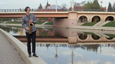 genç adam saksafon çalıyor. Müzisyen parapet kamera, kamera için harekete geçti yürür Nefesli/Üflemeli Saz üzerinde oynar. Adam dar yolda taşır. adam arka plan şehrin üzerinde