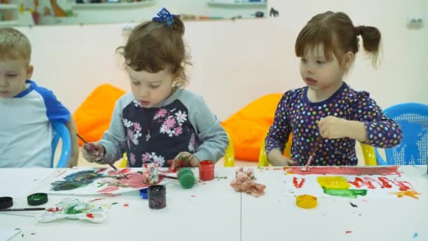 Niños desarrollando una sala de juegos. Emociones de los niños pequeños durante las clases de entretenimiento. niños pintan con pinturas de dedos en hojas blancas de papel. niños estiran sus manos en la pintura untada — Vídeos de Stock