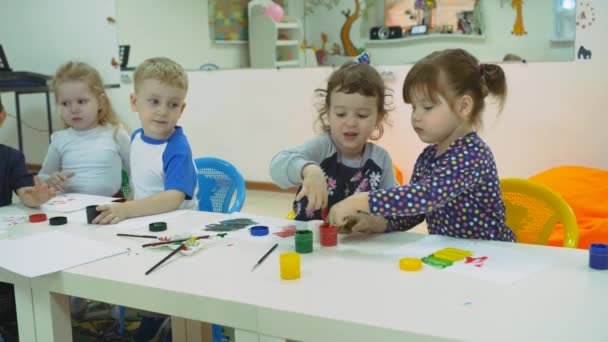 ゲーム ルームの開発子供。クラスを接待中に幼児の感情。子供たちは、紙の白いシートに指塗料でペイントします。子供は塗ったペイントで彼らの手を伸ばす — ストック動画