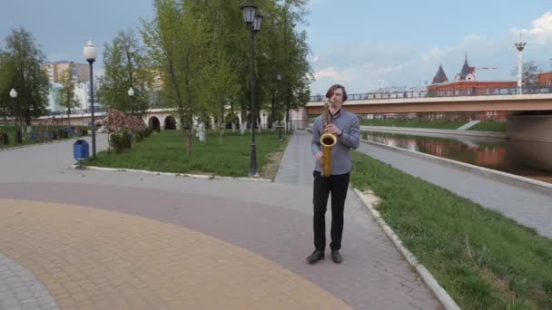 Un tânăr cântă la saxofon. Omul de pe fundalul peisajului orașului suflă în țeavă. Muzicianul merge la camera de-a lungul digului râului . — Videoclip de stoc