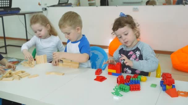 Crianças a desenvolver uma sala de jogos. Emoções de crianças pequenas durante as aulas de entretenimento. Crianças constroem edifícios a partir de figuras geométricas de plástico e madeira . — Vídeo de Stock