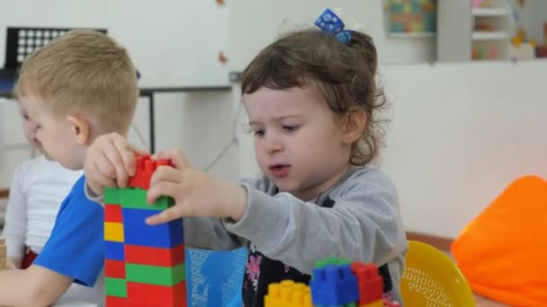 Sala giochi per bambini. Emozioni dei bambini durante le attività ricreative. Ragazzi e ragazze giocano con blocchi di plastica multicolore, seduti al tavolo. ragazza mette il cubo sul dado . — Video Stock