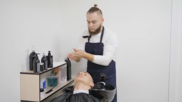 Cabeleireiro para homens. Barbearia. Um jovem tem um corte de cabelo e um serviço de cabeleireiro de um homem barbudo com um cabelo amarrado na cabeça. Lavar a cabeça dos clientes com detergentes. o homem está torcendo a cabeça — Vídeo de Stock