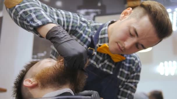 Peluquería para hombres. Barbería. Cuidando la barba. Un peluquero con un corte de pelo trabaja sobre la barba y el bigote de un tipo barbudo. El concepto de un estilo de vida hipster. Cliente Hipster obtiene un — Vídeo de stock