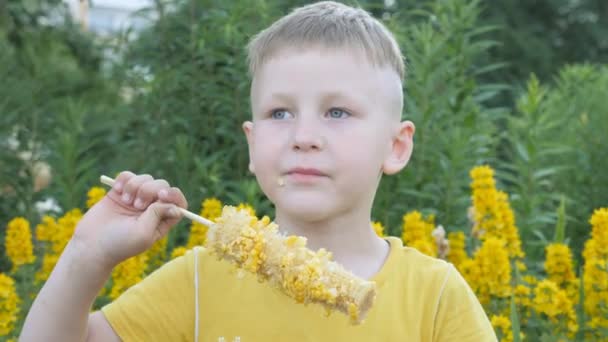 Boy is het eten van maïskolven. kind in een geel T-shirt op een achtergrond van gele bloemen met een eetlust-snacks. — Stockvideo