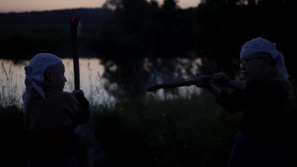 Sparks from burning sticks. Battle of children to burning dubankah. children fight against the background of the river. Coals from burning sticks scatter in different directions. Slow motion — Stock Video