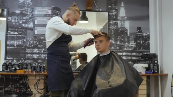 Peluquería para hombres. Barbería. Un joven recibe un corte de pelo y un servicio de cuidado del cabello de un hombre barbudo con un cabello atado a la cabeza. Corte de pelo en el lado de la cabeza de los chicos jóvenes con una cortadora de pelo — Vídeos de Stock