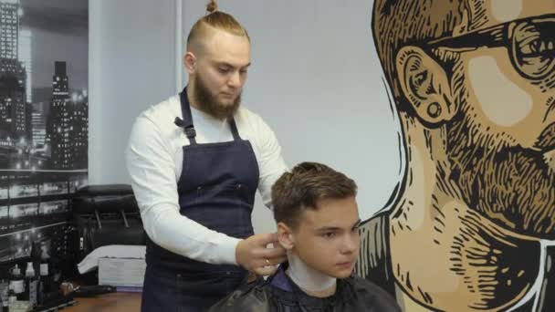 Peluquería para hombres. Barbería. Un joven recibe un corte de pelo y un servicio de cuidado del cabello de un hombre barbudo con un cabello atado a la cabeza. Imposición de un collar desechable blanco en el cuello de los clientes — Vídeos de Stock