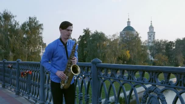 El saxofonista toca la trompeta. City Embankment. un hombre con bigote batido tocando un instrumento musical en las calles de la ciudad. músico se para en el puente y toca un instrumento musical . — Vídeos de Stock