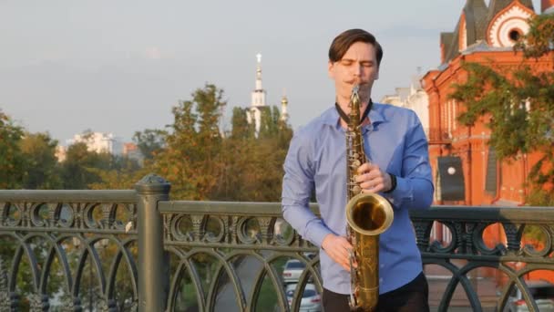 Saxofonista toca trompete. City Embankment. um homem com bigode a tocar um instrumento musical nas ruas da cidade. músico fica na ponte e toca um instrumento musical — Vídeo de Stock