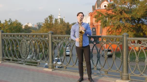 El saxofonista toca la trompeta. City Embankment. un hombre con bigote batido tocando un instrumento musical en las calles de la ciudad. músico se para en el puente y toca un instrumento musical — Vídeos de Stock