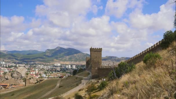 Montagne contro il cielo blu con nuvole bianche. Le nuvole di cirri attraversano il cielo blu. Parte del muro della fortezza sullo sfondo della città situata nella valle. Figure turisti in movimento lungo — Video Stock