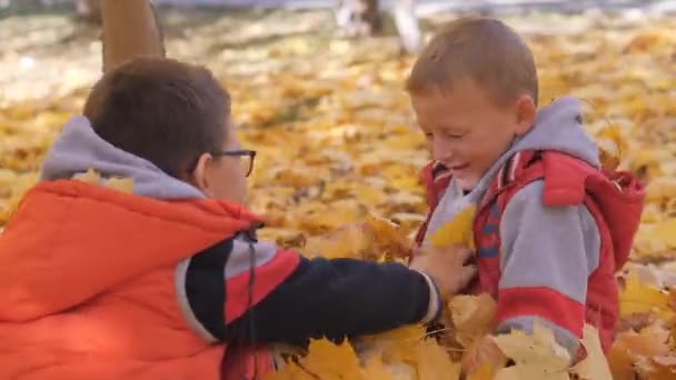 Outono. Crianças pequenas nas folhas amarelas. As crianças brincam na rua com folhas caídas. Árvore de outono de vidoeiros e maçãs. Os rapazes vomitam folhas caídas de árvores no topo. As crianças sentam — Vídeo de Stock