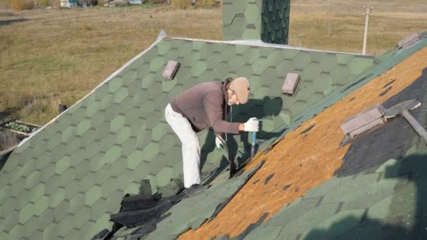 Dismantling the soft roof. French green tile. Roofer working on a sloping roof. A man with a beard tears off an old roofing material from a wooden slab with the help of a crowbar. Construction work at — Stock Video