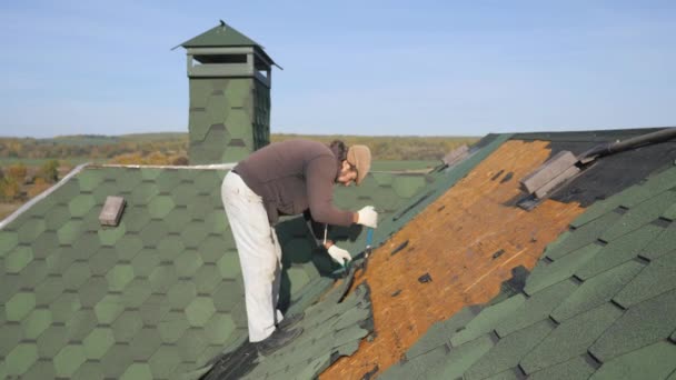 Ontmanteling van het zachte dak. Franse groene tegel. Dakdekker werken op een hellend dak. Een man met een baard tranen uit een oude dakbedekking uit een houten plaat met behulp van een Koevoet. Bouwwerkzaamheden op — Stockvideo
