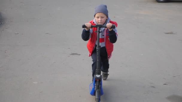 Enfant en scooter. petit garçon pousse avec son pied et monte sur la planche. L'enfant tient la roue et conduit un scooter. tir lent — Video
