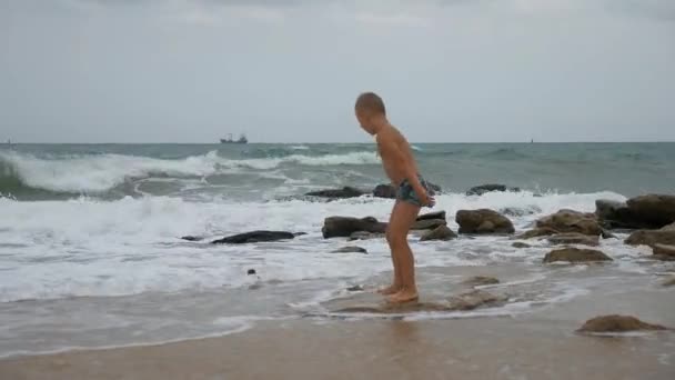 在汹涌的大海中的男孩。穿着泳裤的男孩在强风中站在石头上的时候, 试图保持平衡. — 图库视频影像