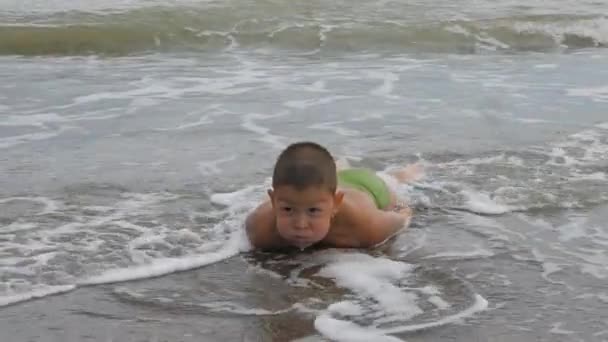 Kind ligt op zijn buik op de zanderige oever in de golven van de zee. jongen laat de fontein van zout water uit zijn mond. Kid gewassen zee golven. — Stockvideo