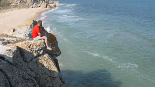 Man Sitter Kanten Klippa Vågorna Tvätta Den Sandiga Stranden Ridge — Stockvideo