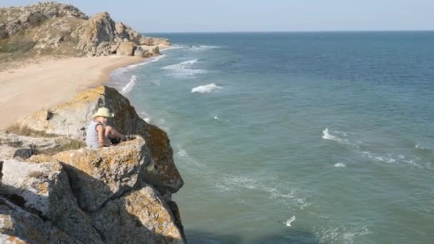 Pojken sitter på kanten av en klippa. Vågorna tvätta den sandiga stranden. Ridge är bruten på klipporna — Stockvideo
