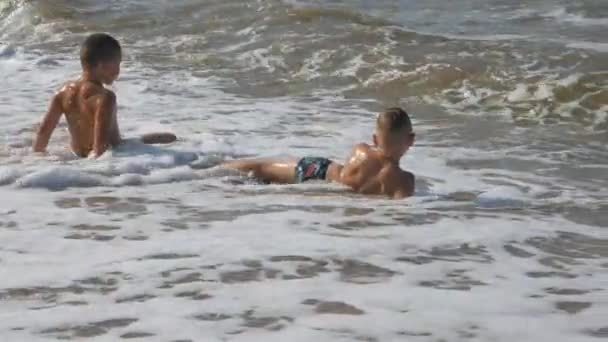 Ребенок на пляже, в морской волне. Два мальчика на золотом песке играют в серфинге. махать головой, прикрывая ребенка . — стоковое видео