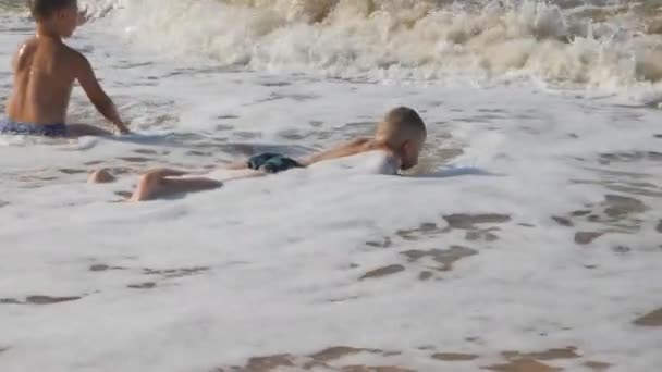 Дитина на пляжі, на морській хвилі. Два хлопці на золотому піску грають у серфінгу. хвиля з головою покриває дитину . — стокове відео