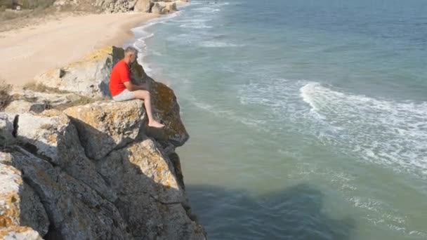 Man Sitter Kanten Klippa Vågorna Tvätta Den Sandiga Stranden Ridge — Stockvideo