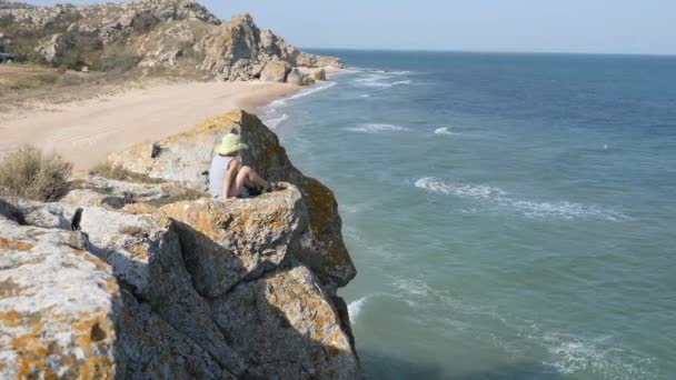 El chico se sienta en el borde del acantilado. Las olas lavan la orilla arenosa. Ridge está roto en las rocas — Vídeos de Stock