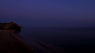Şafak vakti. Deniz dalgaları kumlu plajı yıkıyor. Kayalık bir uçurum denize doğru çıkıntı. Gökyüzü renk değiştirir.