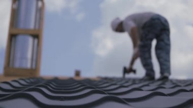 Çatı tamiratı işe yarar. Kurucu, metali çatıdaki sandığa vidalıyor. Çatı inşaatı. Özel bir evin çatısının yarısı metalle kaplıdır. Bulanık elbiseli adam. Bulutlu mavi gökyüzüne karşı bulanık bir figür..
