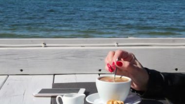 young woman in black leather jacket stirring coffee with a teaspoon in an outdoor cafe near the sea on sunny day. hand closeup