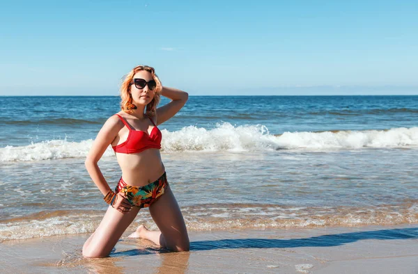 若いブロンドの女の子です赤い水着とサングラス立っています海岸に彼女の手で彼女の頭の後ろ晴れた夏の日に — ストック写真