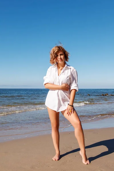 白い水着に身を包んだ若い金髪の笑顔の女性と夏の晴れた日に海岸に立つシャツ — ストック写真