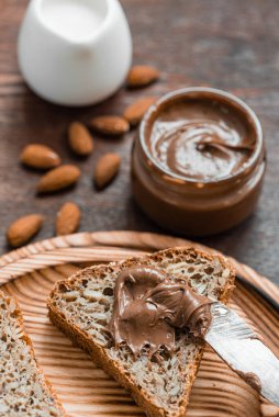Lezzetli bir kahvaltı. Çikolatalı fındık ezmeli cam kavanoz, tahıl ekmeği ve tahtadan bir masada badem..