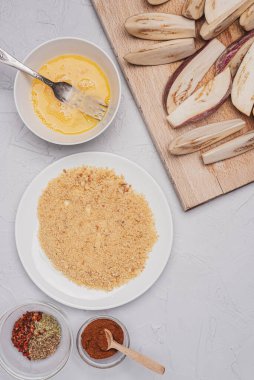 Gri bir yüzeyde dilimlenmiş taze patlıcan, baharat, ekmek kırıntıları ve çırpılmış yumurta..