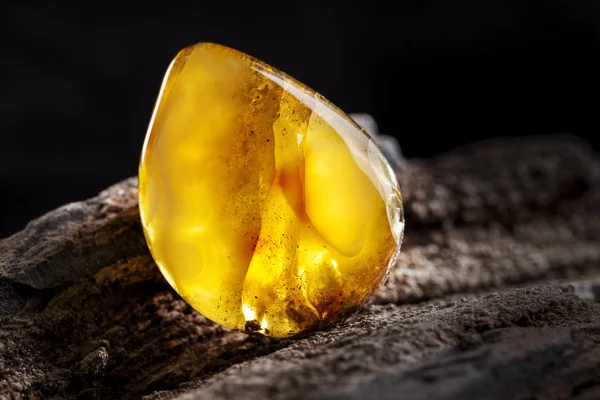 Natural amber. A piece of yellow opaque natural amber on large piece of dark stoned wood. — Stock Photo, Image