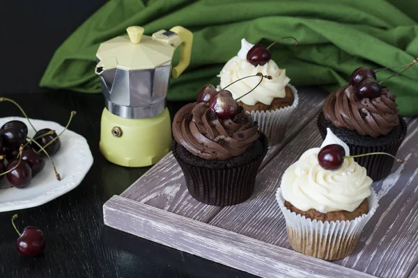 Vanilla cupcakes and chocolate cupcakes with cherries, a plate of berries and a coffee pot on a dark background