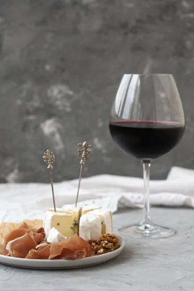 水晶酒杯 红葡萄酒 前景是一个盘子与火腿 饼干和卡门贝尔奶酪 灰色混凝土背景上的成分 — 图库照片