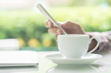 Erkek eller akıllı telefon ve kahve fincanını açık havada, bulanık yeşil bitki arka planında tutuyor.