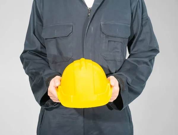 灰色の背景に隔離された黄色の硬い帽子を保持する青いカバーオールに立つ労働者 — ストック写真