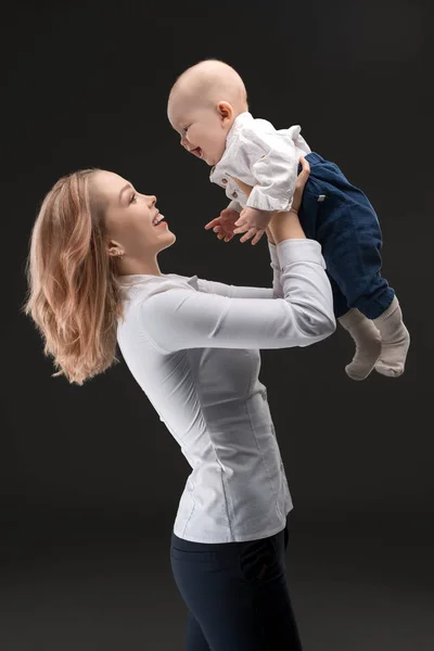 黒の背景に白いシャツとジーンズの彼女持って着てかなり幸せな女小さな赤ちゃん男の子プロフィール撮影 — ストック写真