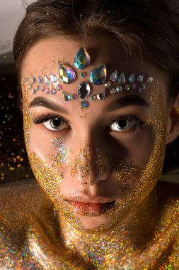 Genç bir kadın, alnına elmas elmaslar yapıştırılmış ve yüzünde ve göğsünde altın parıltısıyla izleyiciye bakıyor..