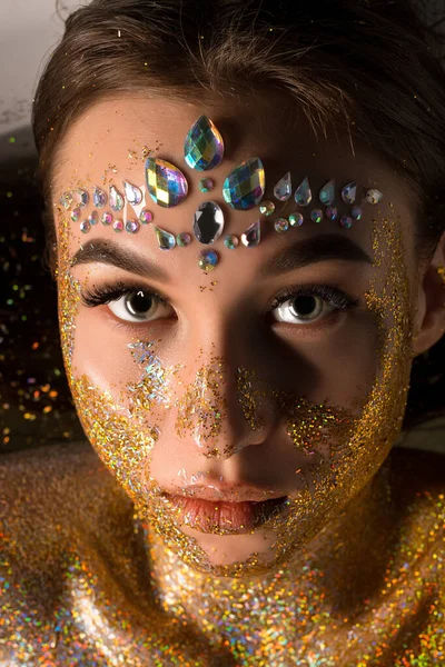 Genç bir kadın, alnına elmas elmaslar yapıştırılmış ve yüzünde ve göğsünde altın parıltısıyla izleyiciye bakıyor..