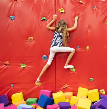 Küçük gülümseyen sarışın kız oyun odasında kırmızı tırmanma duvarının yakınında ayakta