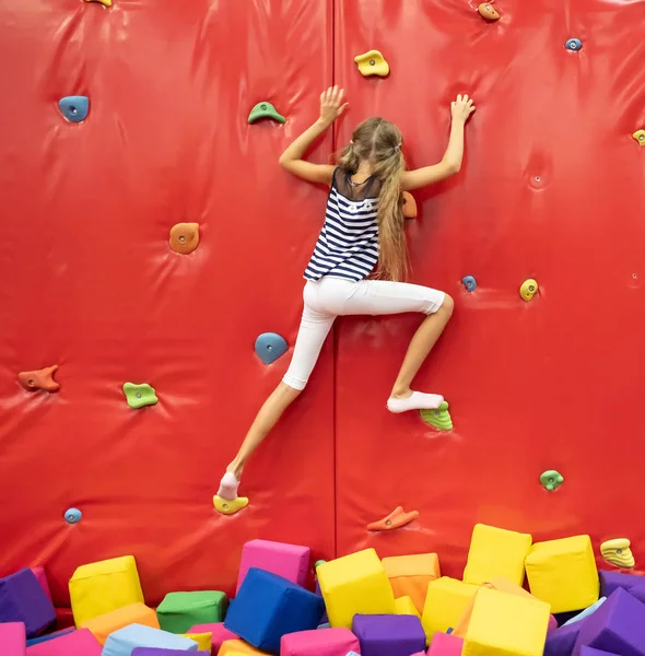 Pequena menina loira sorridente de pé perto da parede de escalada vermelha na sala de jogos — Fotografia de Stock