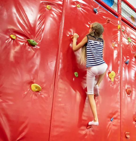 Pequena menina loira sorridente de pé perto da parede de escalada vermelha na sala de jogos — Fotografia de Stock