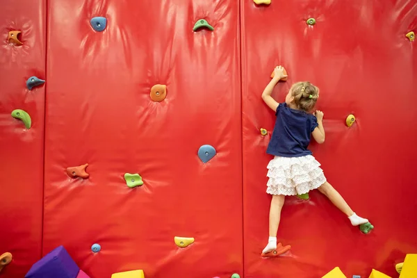 Pequeña chica rubia sonriente de pie cerca de la pared de escalada roja en la sala de juegos — Foto de Stock
