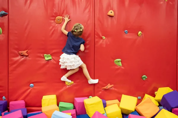 Little smiling blond girl standing near red climbing wall in the play room — Stock Photo, Image
